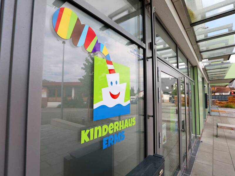Der Eingang eines modernen Gebäudes mit Glasfassade zeigt ein buntes Logo und den Schriftzug "Kinderhaus Erms".