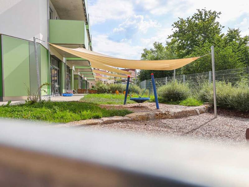 Ein moderner Spielplatz mit Schaukel und Sonnensegeln vor einem Gebäude mit grünen Fassaden.