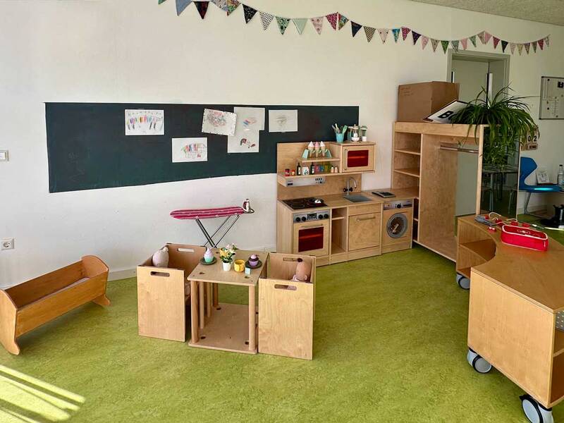Das Bild zeigt einen hellen Kindergarten- oder Klassenraum mit Spielküche, kleinen Holzmöbeln, Kinderzeichnungen an der Wand und grünem Teppichboden.