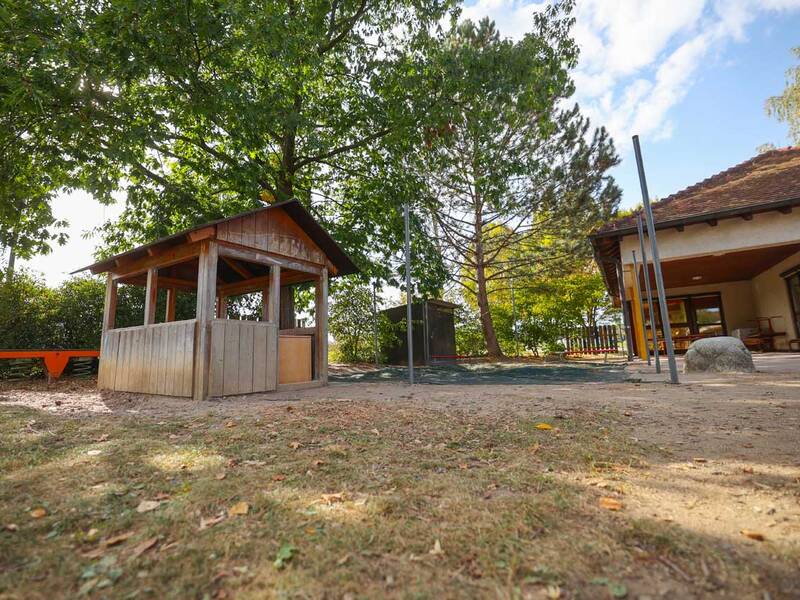Ein Spielplatz mit einem kleinen Holzhaus und einer Wippe, umgeben von Bäumen, befindet sich neben einem Gebäude mit großem Vordach.
