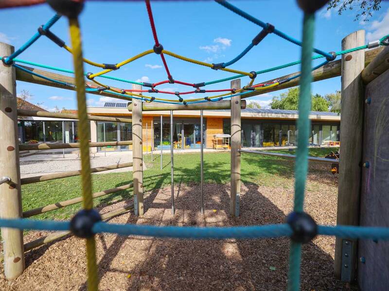 Ein buntes Klettergerüst aus Seilen und Holzstangen steht auf einem Spielplatz vor einem modernen Gebäude mit viel Glas.
