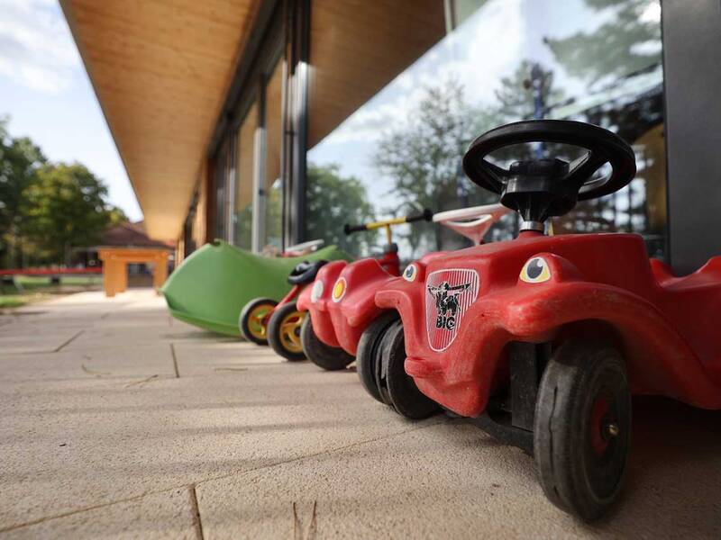 Mehrere rote Kinderrutschautos stehen nebeneinander auf einer gepflasterten Fläche vor einem Gebäude mit Glasfront, umgeben von einem grünen Außenbereich.