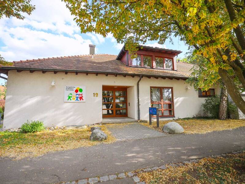Ein modernes, eingeschossiges Gebäude mit rotem Dach und Holzelementen, umgeben von herbstlich gefärbten Bäumen, beherbergt einen evangelischen Hort, erkennbar an einem Schild neben dem Eingang.