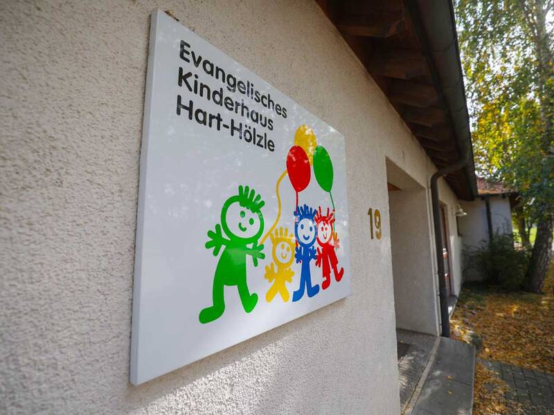 Ein Schild mit der Aufschrift "Evangelisches Kinderhaus Hart-Hölzle" zeigt bunte Kindergrafiken mit Luftballons, montiert an einer Hauswand.