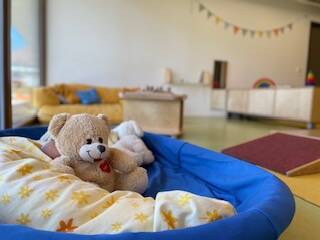 Ein Stoffteddybär sitzt in einem blauen, mit einer Decke ausgekleideten Korb in einem hellen, kindgerechten Raum mit gelbem Sofa und bunten Girlanden im Hintergrund.