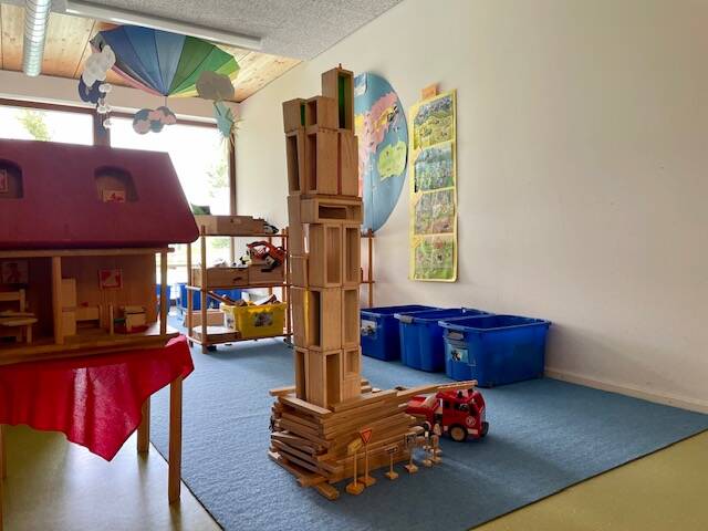 In einem hellen Raum mit blauem Teppichboden steht ein hoher Turm aus Holzklötzen, umgeben von Spielzeug und blauen Kisten, während im Hintergrund eine bunte Dekoration von der Decke hängt.