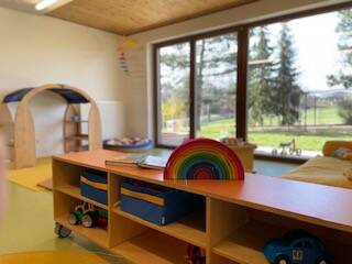 Ein heller Kindergartenraum mit Spielzeugregalen und Spielzeugautos vor großen Fenstern, die einen Blick auf einen grünen Außenbereich bieten.