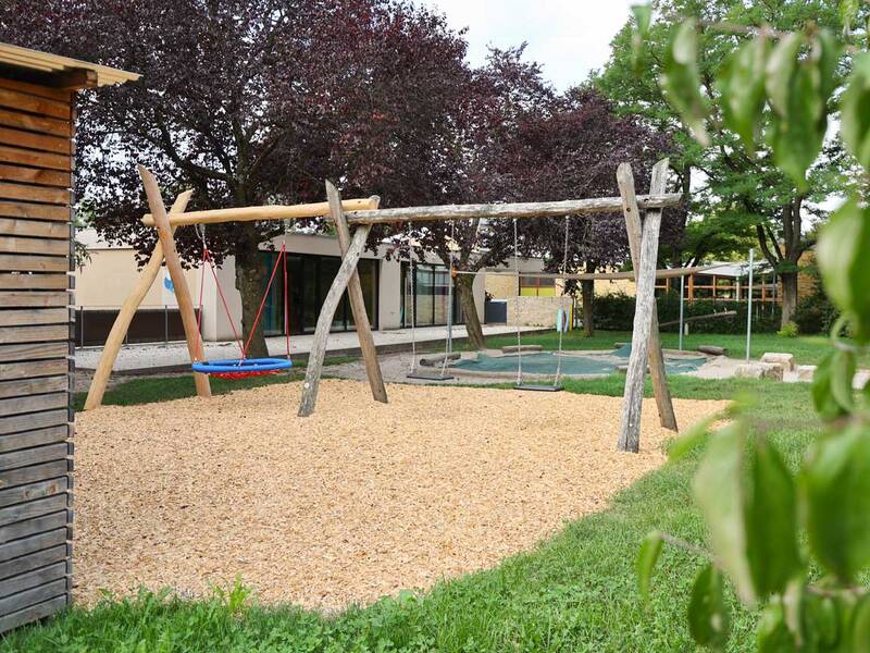 Ein Spielplatz mit einer Holzschaukel steht auf einem Untergrund aus Holzschnitzeln neben einem Holzschuppen, umgeben von grünen Bäumen und Büschen.