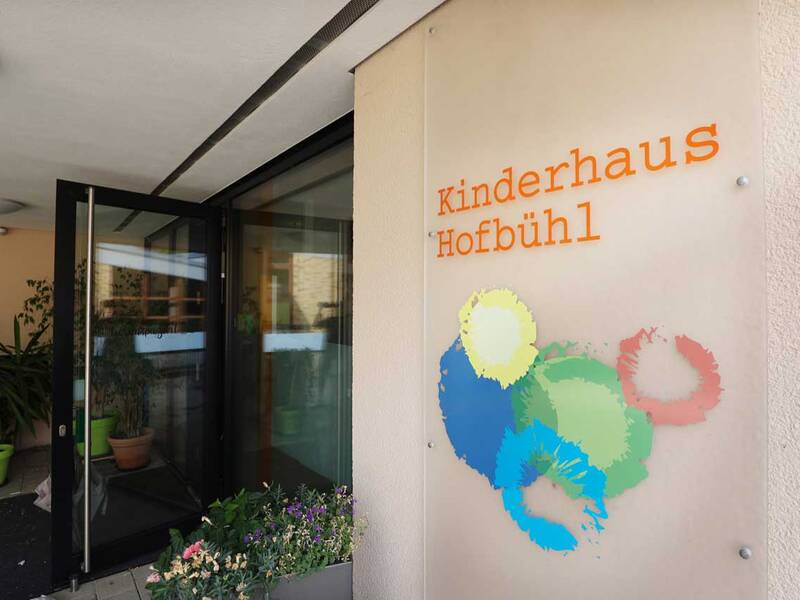 Das Bild zeigt den Eingang eines Gebäudes mit der Beschriftung "Kinderhaus Hofbühl" und bunten Farbtupfern auf einem Schild, sowie Pflanzen im Hintergrund.