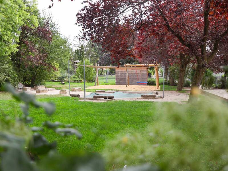 Ein ruhiger Spielplatz mit grüner Wiese, umgeben von Bäumen, bietet Schaukeln und Kletterstrukturen aus Holz.