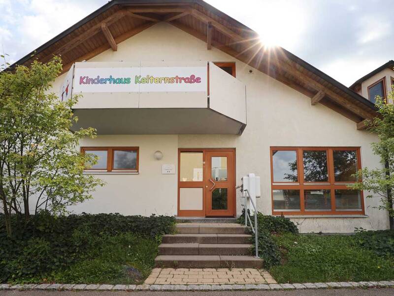 Das Bild zeigt die Fassade eines Gebäudes mit Holzdach und großen Fenstern, über dessen Eingang ein Schild mit der Aufschrift "Kinderhaus Kelternstraße" hängt, während die Sonne von hinten scheint.