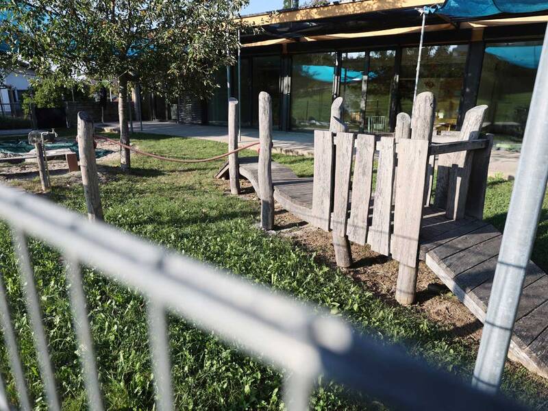 Ein moderner Spielplatz mit Holzelementen und einem Seilhindernis befindet sich vor einem Gebäude mit Glasfront.