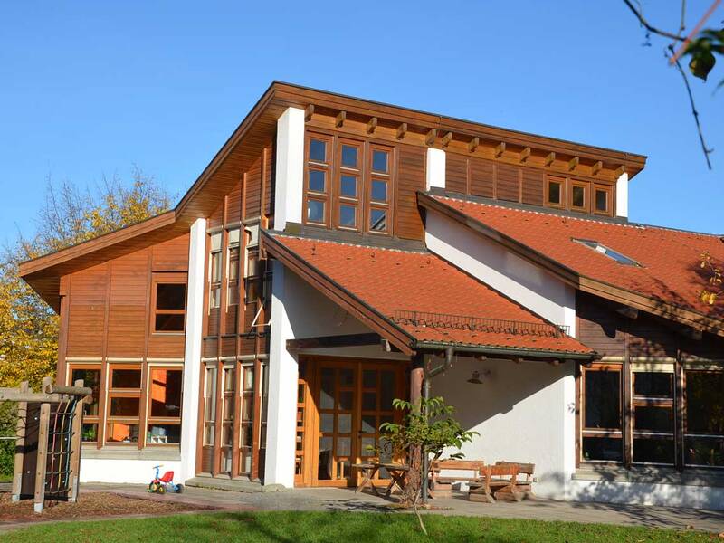Ein modernes Holzhaus mit rotem Schrägdach steht in einer grünen Umgebung unter einem klaren blauen Himmel.