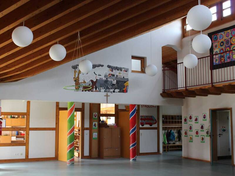Ein Innenraum mit Holzdecke und runden Hängeleuchten, der bunt dekoriert ist, darunter eine bemalte Säule und Kinderbilder an den Wänden.