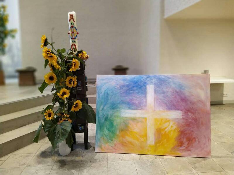 In einem Kirchenraum stehen eine große Osterkerze mit Sonnenblumen geschmückt und ein farbenfrohes Bild mit einem weißen Kreuz.