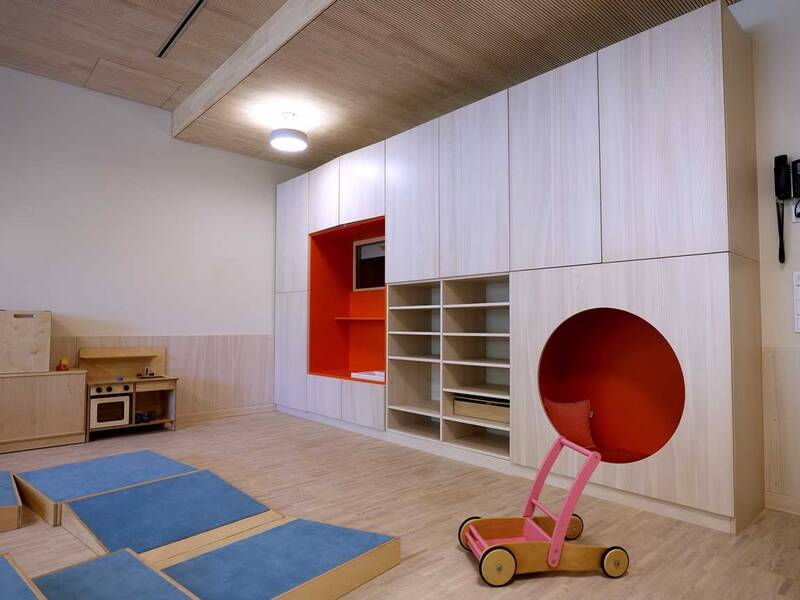 Ein heller Raum mit Holzfußboden und -decke, einer großen Schrankwand mit roten Details, einem kleinen Spielküchenbereich und einem rosa-rote Lauflernwagen auf dem Boden.