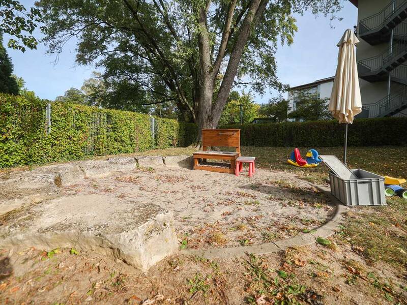 Ein Sandkasten im Freien, umgeben von Bäumen und Sträuchern, mit einem Holzspieltisch, bunten Kinderspielzeugen und einem Sonnenschirm.