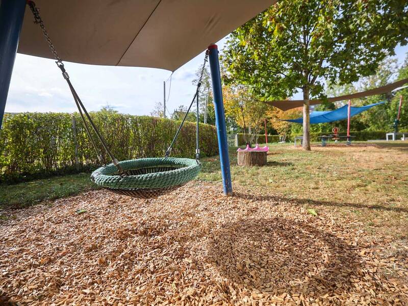 Ein Spielplatz mit einer Nestschaukel im Vordergrund und schattenspendenden Tüchern sowie Bäumen im Hintergrund.