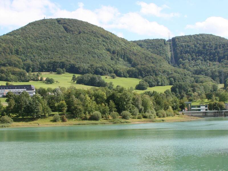 Eine grüne, bewaldete Landschaft mit Hügeln und einem ruhigen See im Vordergrund unter einem blauen Himmel.