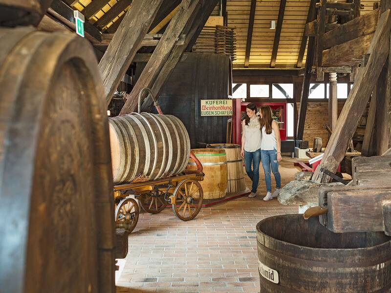 Zwei Personen stehen in einer traditionellen Weinpresse mit großen Holzfässern und Holzbalken.