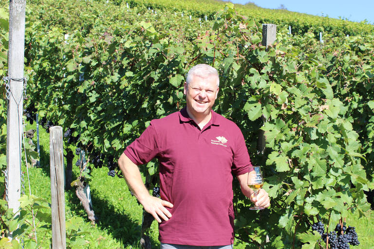 Bruno Seitz Ein Mann in einem weinroten Hemd steht in einem Weinberg und hält ein Weinglas in der Hand.