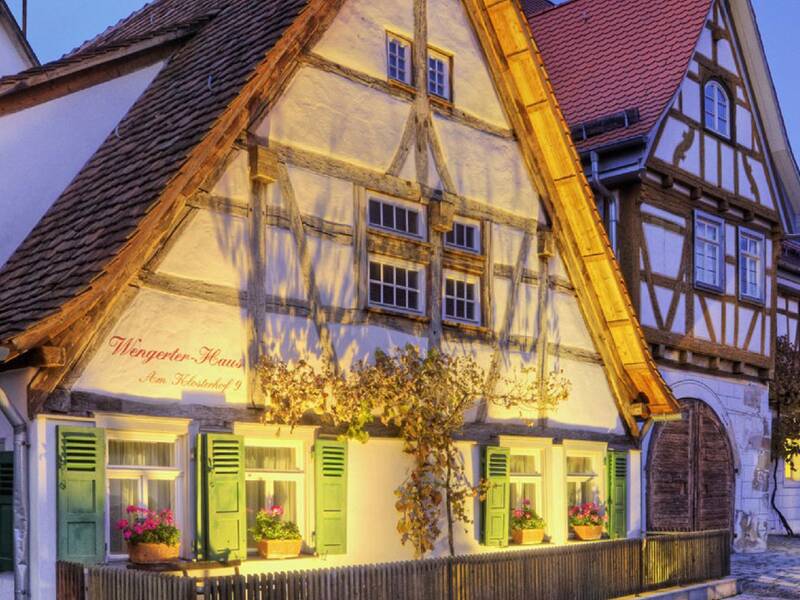 Ein traditionelles, beleuchtetes Fachwerkhaus mit Blumen im Fenster steht in einer gepflasterten Straße bei Dämmerung.