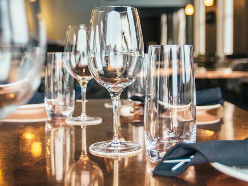 In dem Bild ist ein elegant gedeckter Tisch in einem Restaurant zu sehen, mit mehreren Weingläsern, Tellern und schwarzen Stoffservietten.