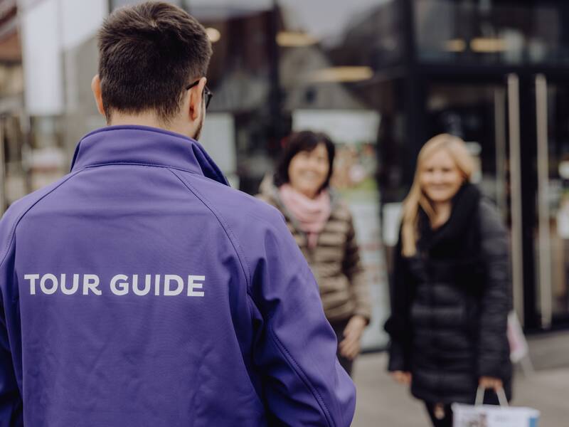 Ein Tourguide in einer lilafarbenen Jacke steht vor zwei lächelnden Frauen, die am Eingang eines Gebäudes stehen.