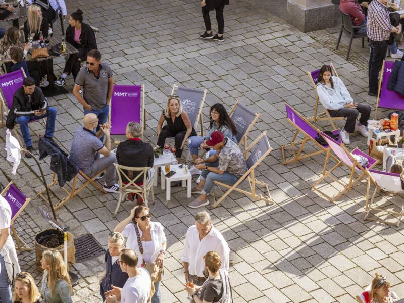 Menschen sitzen und stehen auf einem gepflasterten Platz vor einem Café, einige entspannen auf Liegestühlen und andere plaudern in Gruppen.