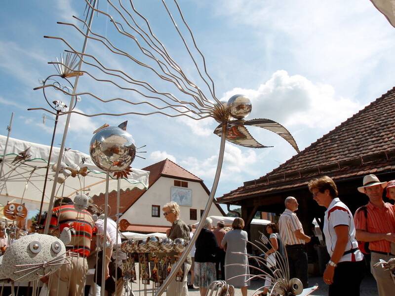 Auf einem sonnigen Kunsthandwerksmarkt betrachten Menschen verschiedene skulpturale Kunstwerke aus Metall.