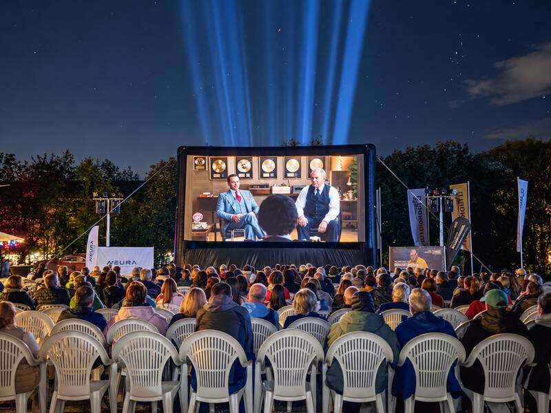 Eine Menschenmenge sitzt in weißen Plastikstühlen und schaut einen Film auf einer großen Leinwand unter einem sternenklaren Nachthimmel.
