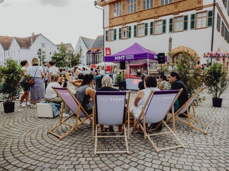 Eine Gruppe von Menschen sitzt auf einem gepflasterten Platz in Liegestühlen vor einem lilafarbenen Zelt mit der Aufschrift "MMT", umgeben von historischen Gebäuden und Passanten.