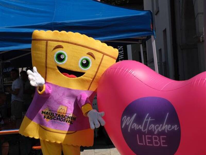 Ein Maskottchen in Form einer Maultasche posiert fröhlich neben einem großen, pinken Herzballon mit der Aufschrift "Maultaschen Liebe" vor einem blauen Pavillon.