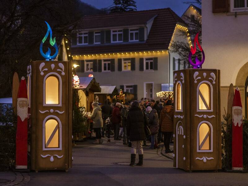 Eine festlich beleuchtete Weihnachtsmarktszene mit dekorativen Leuchtkerzen und Menschen, die entlang von Holzbuden schlendern.