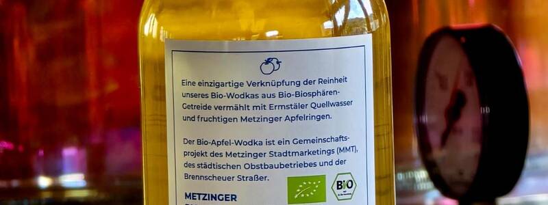 Eine Flasche Metzinger Bio-Apfel-Wodka mit einem detaillierten Etikett vor einem unscharfen, metallischen Hintergrund.