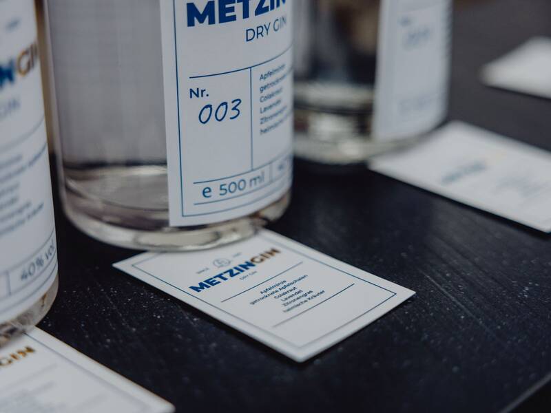 Einige Flaschen mit der Aufschrift "Metz Gin" stehen auf einem dunklen Tisch, begleitet von gleichartigen Informationskarten.