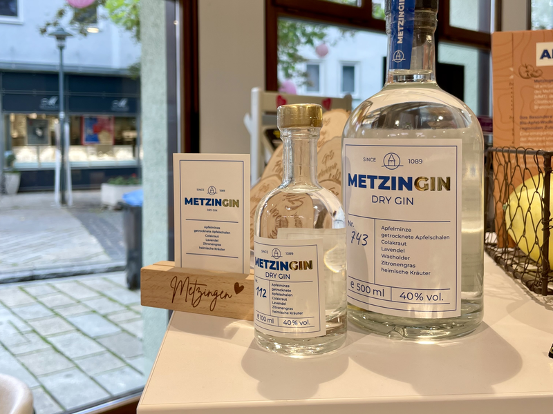 Zwei Flaschen Metzingin Dry Gin stehen auf einem Tisch, umgeben von Fenstern, die einen Blick auf eine Straße bieten.