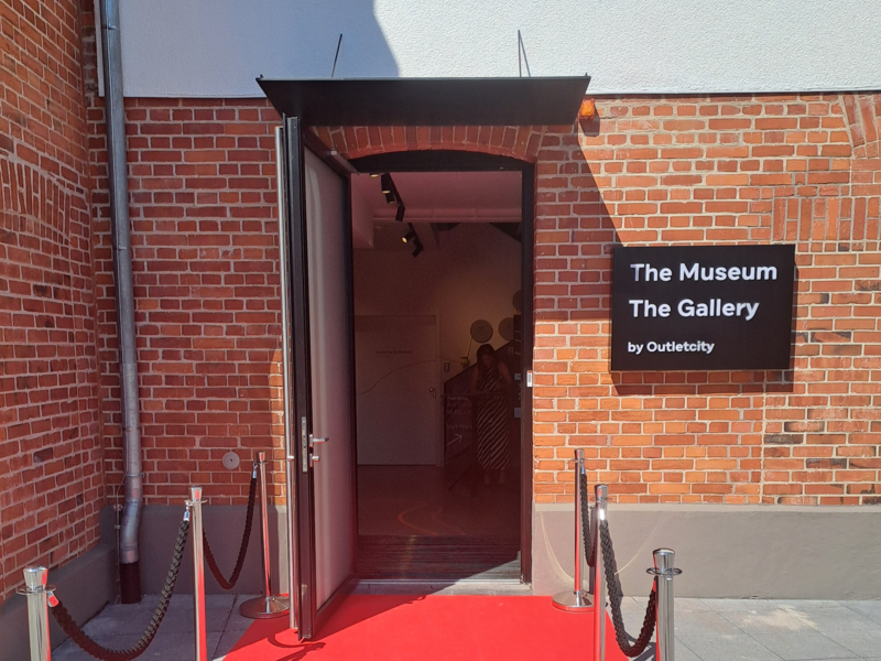 Ein roter Teppich führt zu einem Eingang eines Gebäudes aus roten Ziegelsteinen, auf dem ein Schild mit der Aufschrift "The Museum The Gallery by Outletcity" hängt.