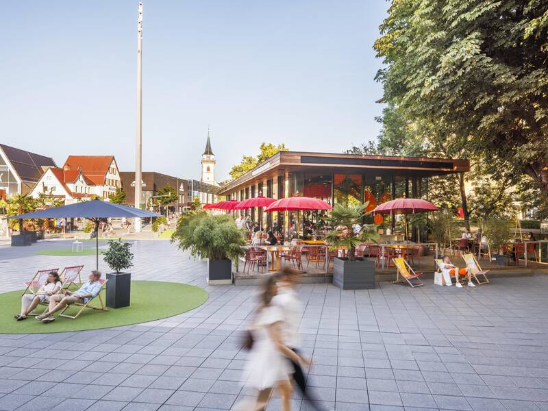 Ein moderner Platz mit Sonnenschirmen, Liegestühlen und einem Café, umgeben von Bäumen und traditioneller Architektur im Hintergrund.
