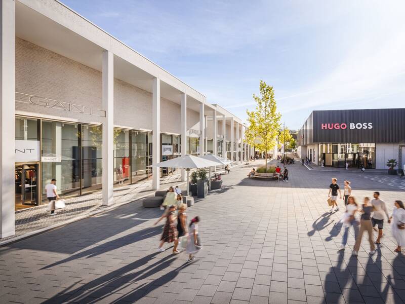 Eine belebte, moderne Einkaufsstraße mit Geschäften von GANT und Hugo Boss und mehreren Personen, die flanieren oder sitzen.
