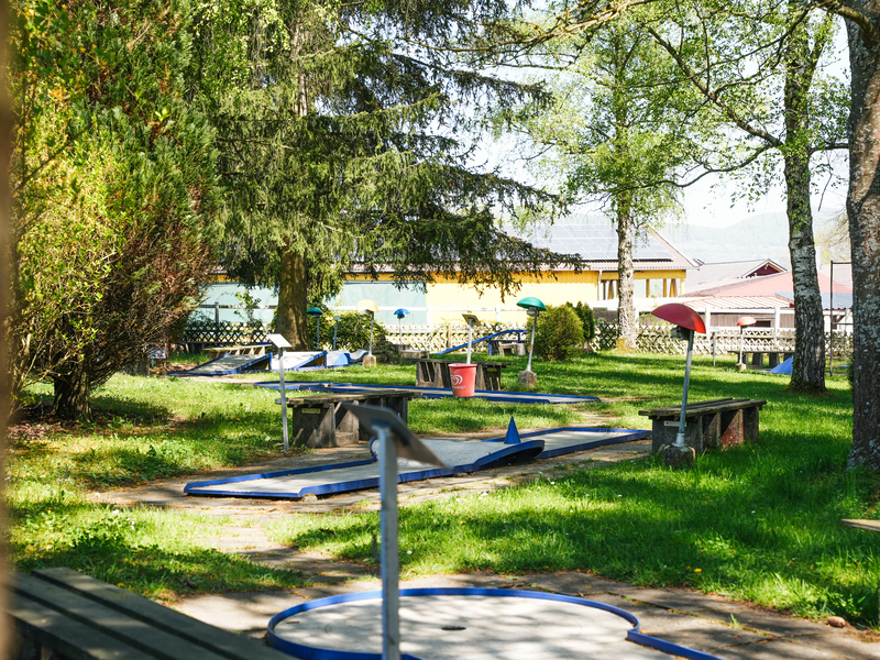 Eine Minigolf-Anlage mit verschiedenen Bahnen und bunten Hindernissen befindet sich in einem parkähnlichen, grünen Umfeld mit Bäumen im Hintergrund.