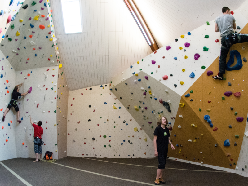 Drei Personen klettern an den Wänden einer Kletterhalle mit bunten Griffen, während eine weitere Person auf dem Boden steht und zuschaut.