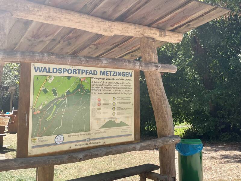 Ein Holzunterstand mit einer Infotafel zum Waldsportpfad Metzingen steht neben einem Abfalleimer, umgeben von Bäumen.