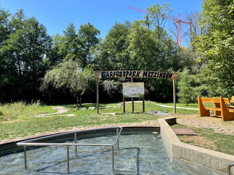 Das Bild zeigt einen Eingang zum Barfußpark Metzingen mit einem Wasserbecken im Vordergrund, umgeben von grüner Natur und einem Holzschild.