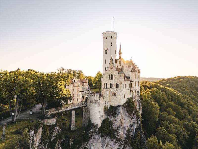 Die Aufnahme zeigt Schloss Lichtenstein, das majestätisch auf einem steilen Felsen thront, von grünen Wäldern umgeben und vom sanften Licht der Abendsonne beleuchtet wird.