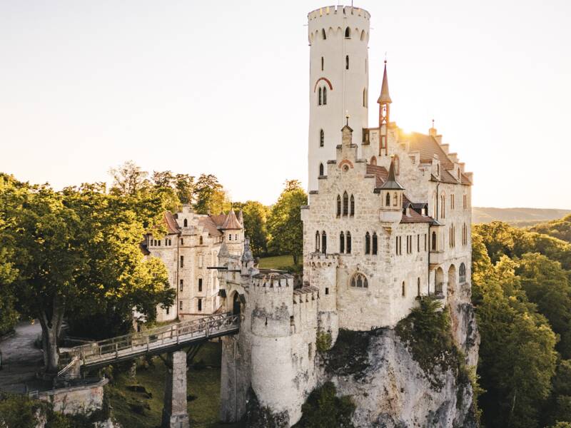 Die Aufnahme zeigt Schloss Lichtenstein, das majestätisch auf einem steilen Felsen thront, von grünen Wäldern umgeben und vom sanften Licht der Abendsonne beleuchtet wird.
