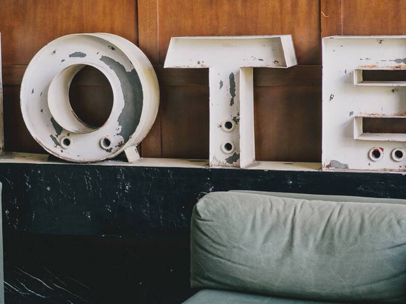 Das Bild zeigt alte, abgenutzte Buchstaben, die das Wort "HOTEL" auf einer dunklen Oberfläche vor einer Holzverkleidung anzeigen, hinter grünen Sofas.