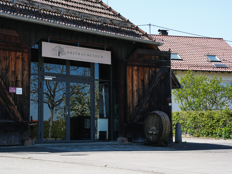 Das Bild zeigt den Eingang eines Gebäudes mit der Aufschrift "Obstbaumuseum", umgeben von Holzstrukturen und Natur, mit einem weiteren Gebäude im Hintergrund.