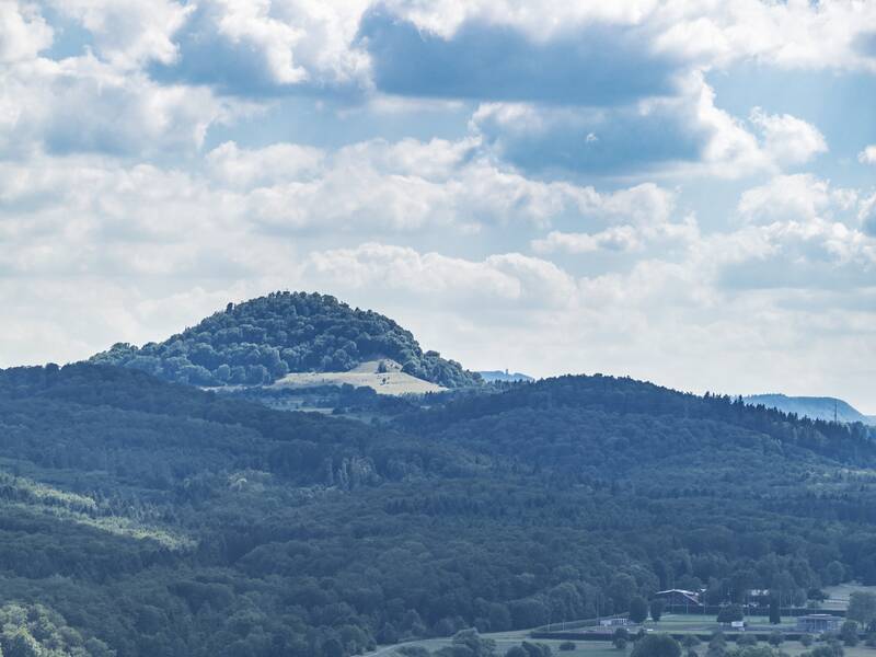 Eine malerische Landschaft mit einem Hügel im Hintergrund, grünen Wiesen und einem kleinen Ort im Vordergrund, unter einem leicht bewölkten Himmel.