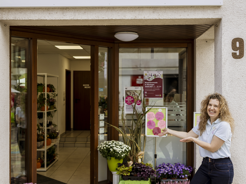 Eine Frau steht lächelnd vor einem Laden namens "florafee café" mit Pflanzen auf dem Boden und einem Willkommensschild.